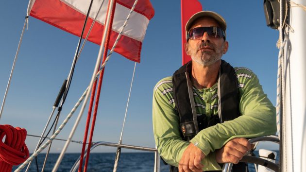 A sailor wearing a lifejacket, sunglasses and a green jumper on his boat
